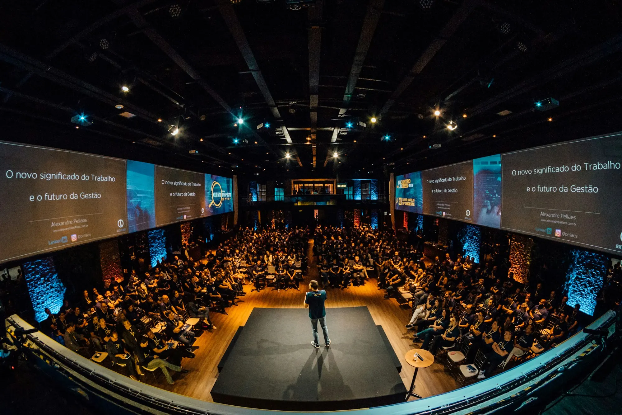 Auditório lotado durante uma convenção, com telas acesas ao redor do palco.